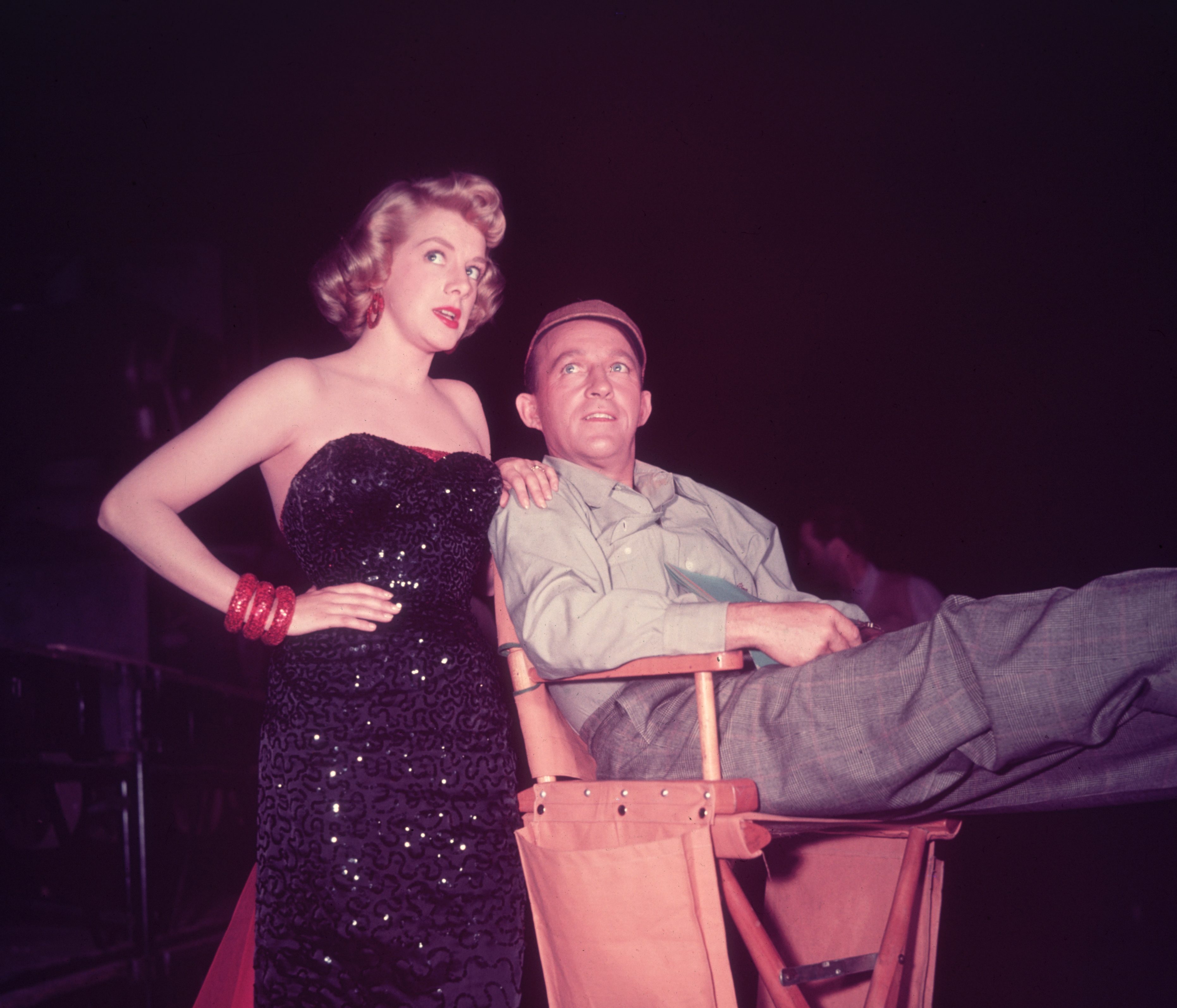 1954: American singer and actors Rosemary Clooney (1928 - 2002) and Bing Crosby (1903 - 1997) talking on the set of director Michael Curtiz's film, 'White Christmas'. Clooney is in costume in a strapless full-length black sequined gown, and Crosby wears a baseball cap and sits in a director's chair. 