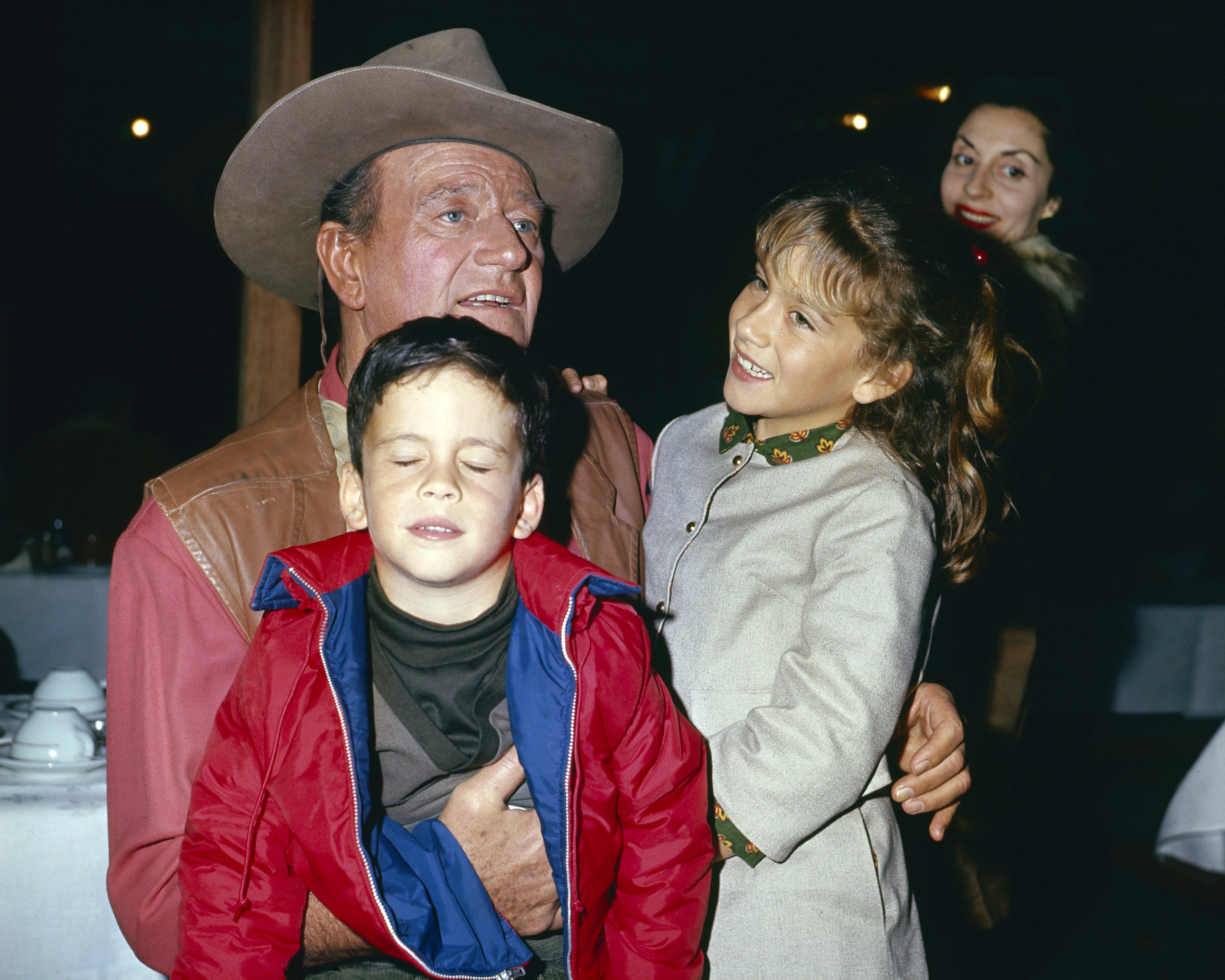 View of American actor John Wayne (1907 - 1979) with two of his children, son John Ethan and daughter Aissa Wayne, and, in the background, his wife, Peruvian-born actress Pilar Pallete, as they are photographed in an unspecified restaurant, 1967. Wayne is in costume (as Cole Thornton) for his role in 'El Dorado' (directed by Howard Hawks). 