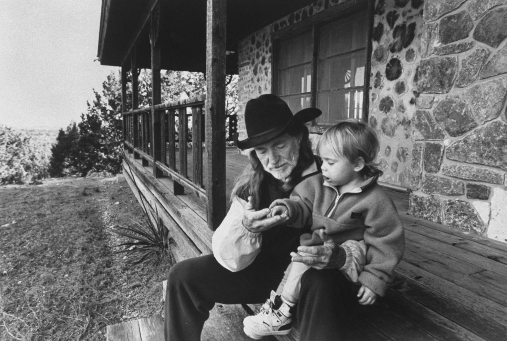 C/W singer Willie Nelson w. his son Lukas Autry, 2, chatting as they sit on front steps of his barren cabin which the IRS stripped of its furniture after it seized the 688-acre property because of his failure to pay back income taxes amounting to 16.7mi. llion