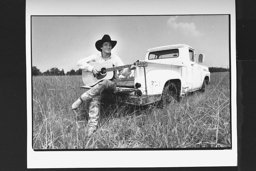 C & W star Clint Black sitting on back of pick-up truck playing guitar in field; near Freedom Fest concert.