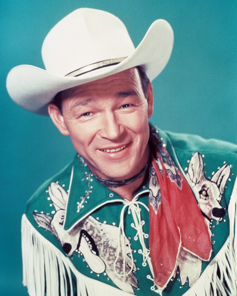 Roy Rogers (1911-1998), US actor and singer, wearing a white cowboy hat, red neckerchief and fringed western shirt in a studio portrait, against a blue background, circa 1950.