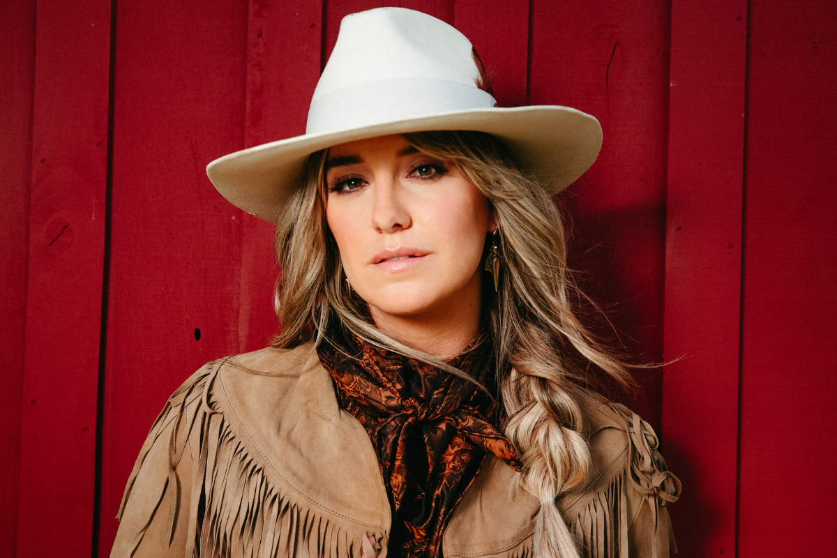 Lainey Wilson poses, wearing white hat and fringe jacket