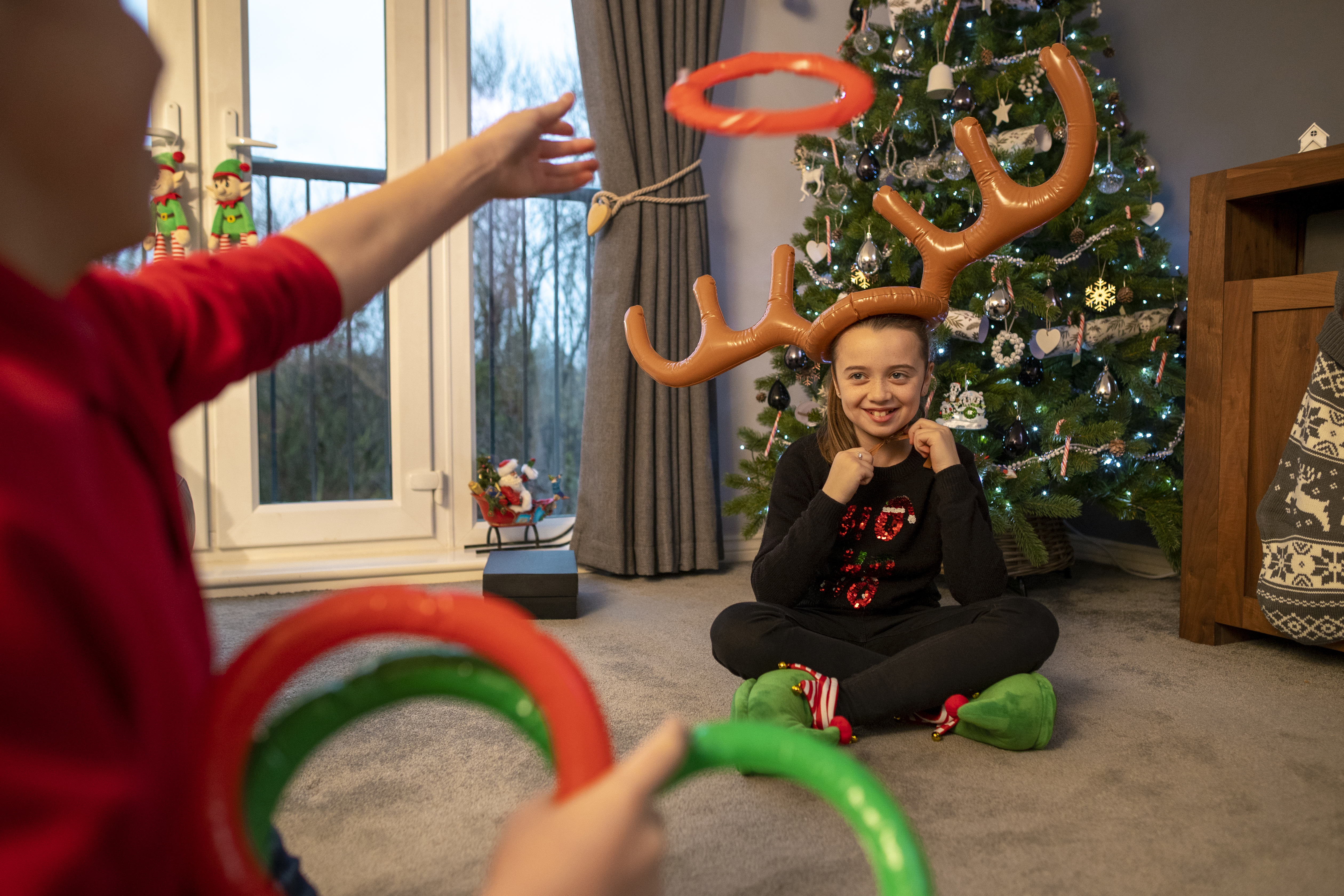 A young girl sitting at home in the living room wearing inflatable reindeer antlers. She is sitting on the floor in the living room at home opposite her brother, who is throwing inflatable rings at the antlers. The room is filled with Christmas decorations, ready for Christmas Day.