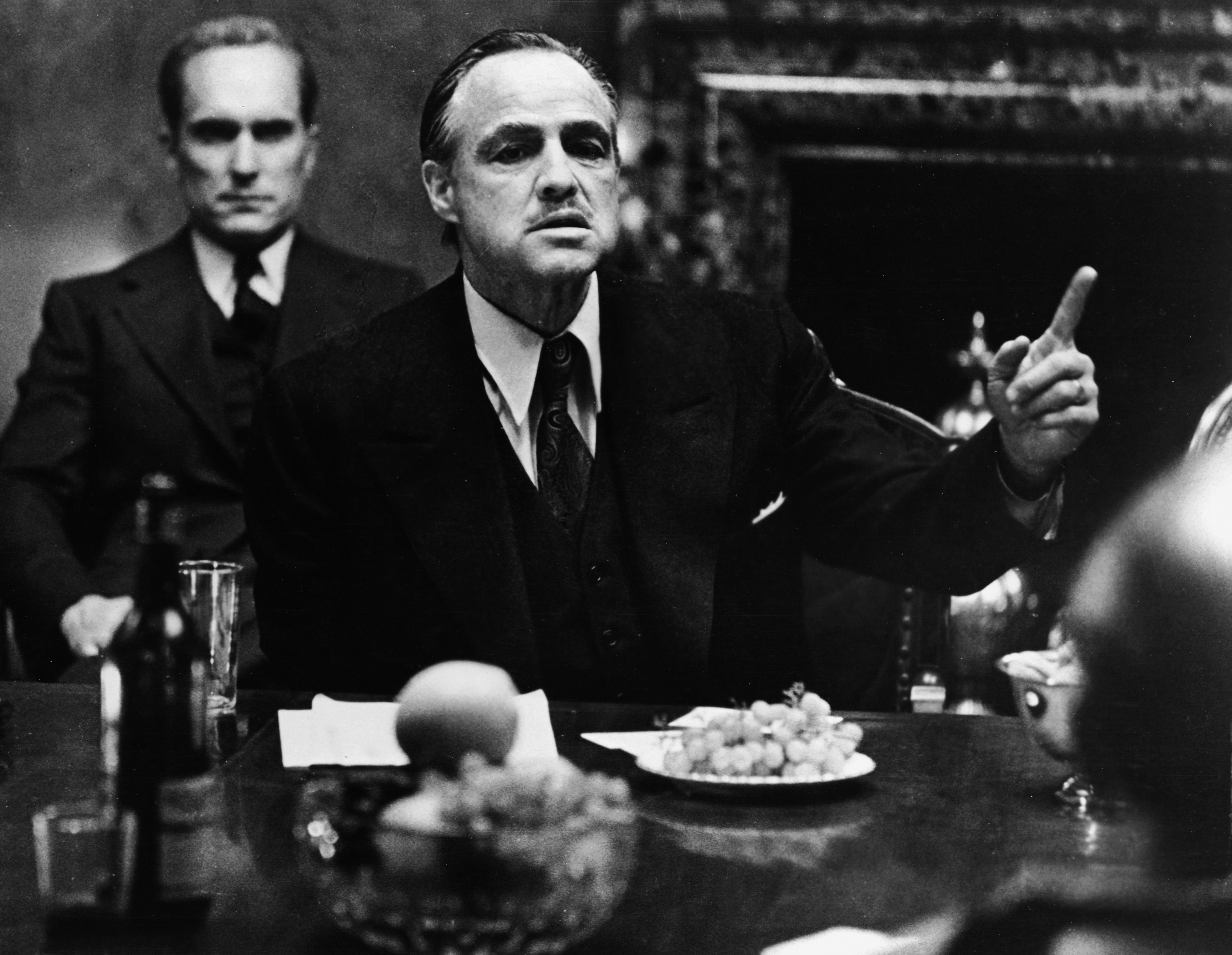 American actor Marlon Brando gestures at a table while American actor Robert Duvall sits behind him in a still from the film, 'The Godfather,' directed by Francis Ford Coppola and based on the novel by Mario Puzo.