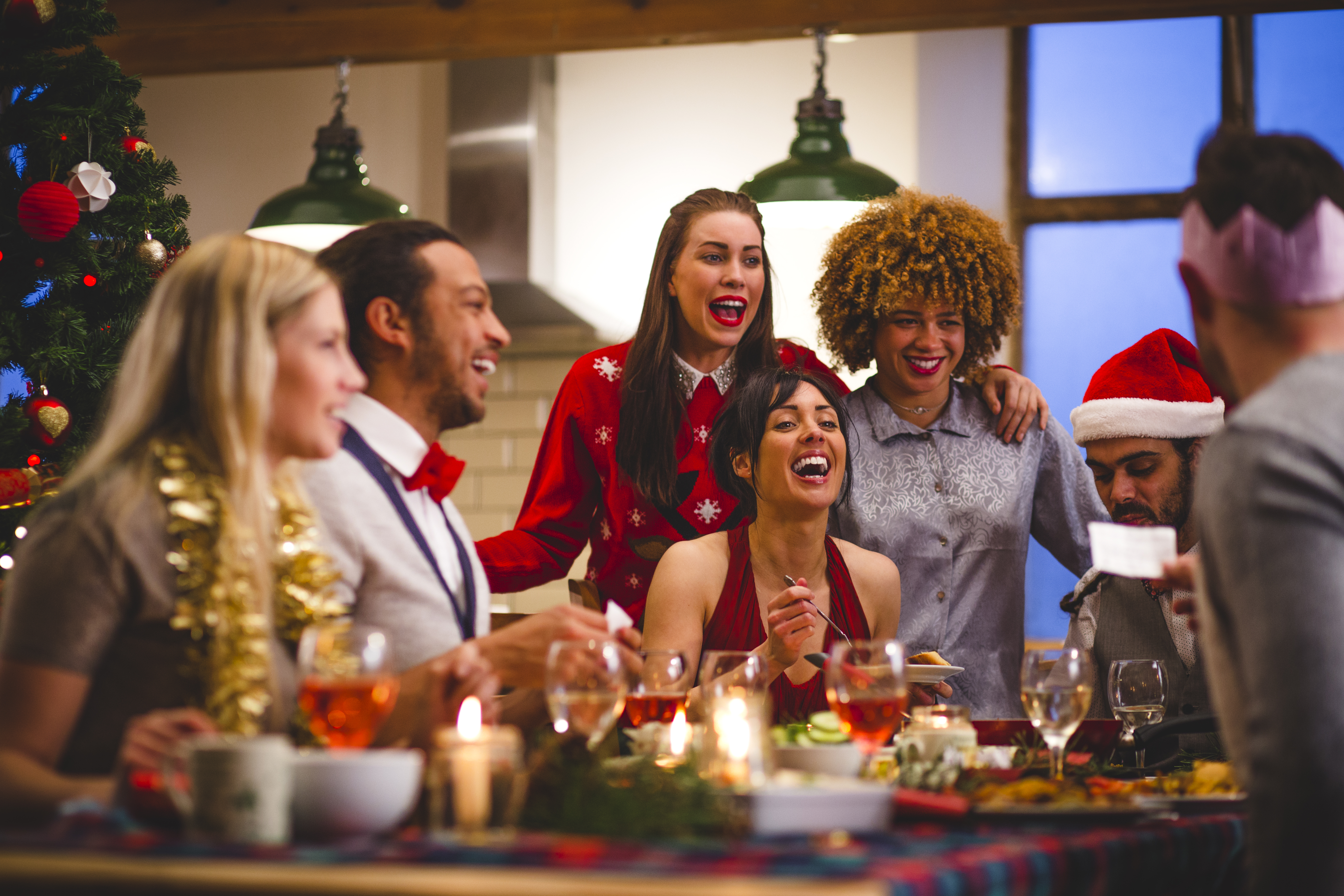 Group of friends laughing as they sit around a table eating and drinking at Christmas. One man is reading a joke from a christmas cracker. Party hats and christmas jumpers are worn.