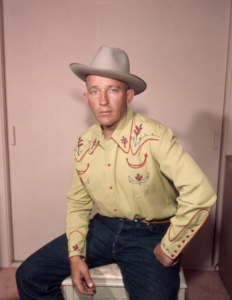 Studio portrait of American actor and singer Bing Crosby seated with his legs spread apart and his fist on his hip, wearing a western shirt, jeans, and a cowboy hat.