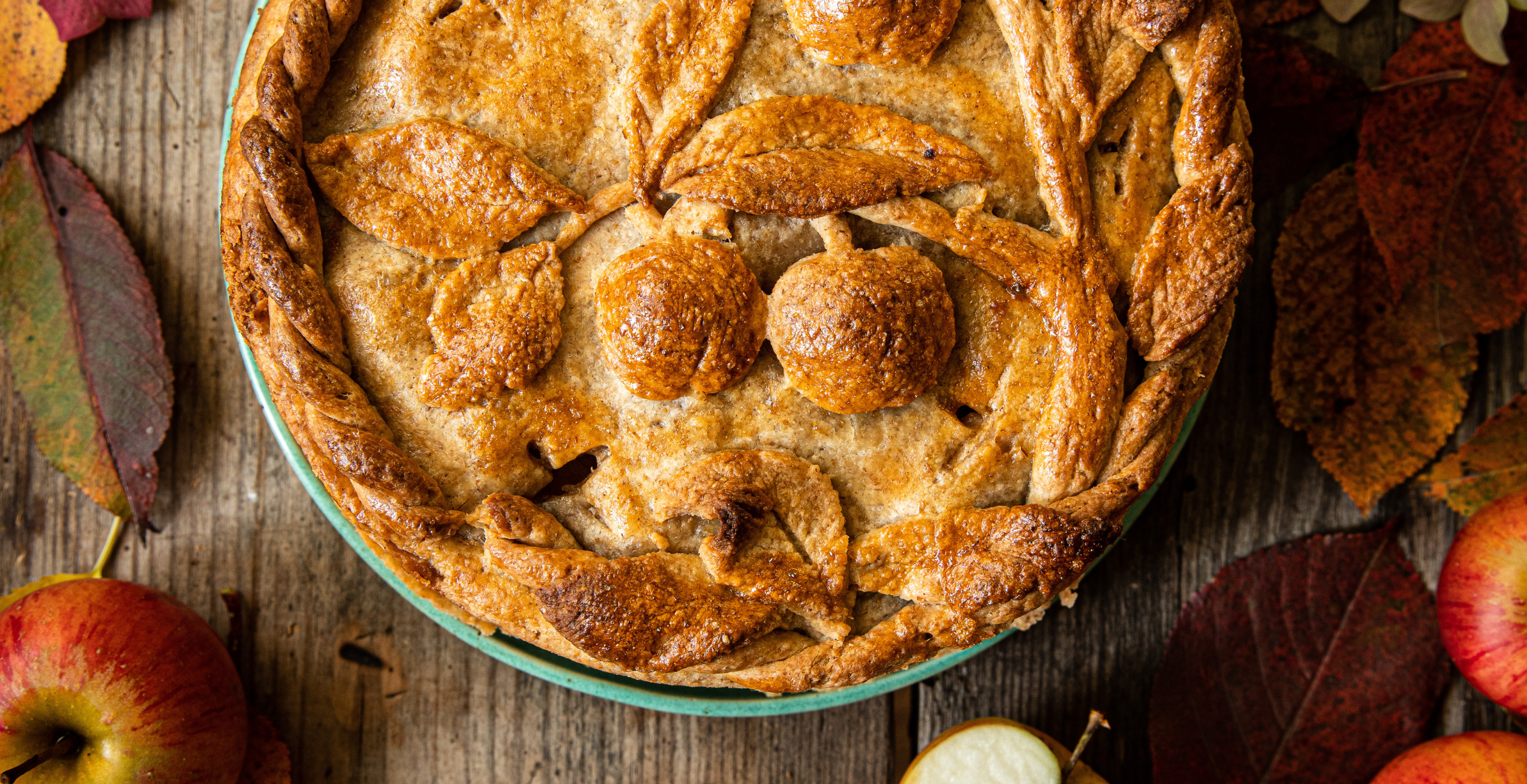 Autumn apple pie with decorated crust surrounded with leaves and red apples on wooden table.