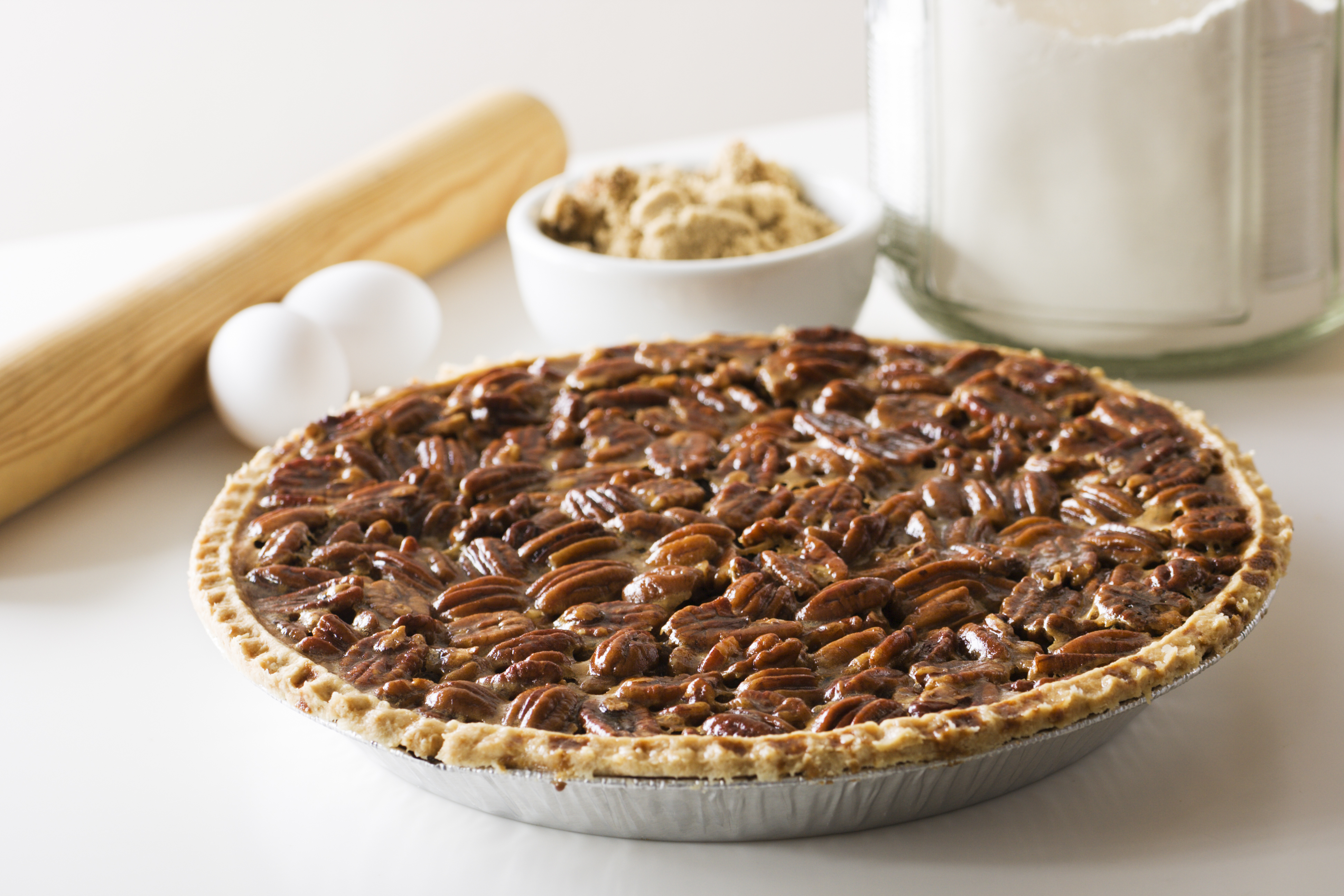 A freshly baked pecan pie with ingredients and a rolling pin in the background. The delicious homemade dessert is ready for a holiday or other special occasion.