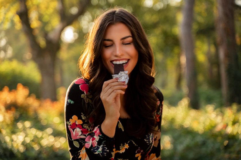 Gorgeous woman eating bar of dark chocolate in the sunny park.